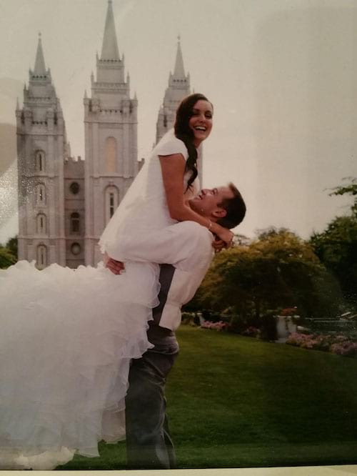 Un homme en tenue de mariage portant sa femme avec une longue robe de mariée blanche devant un château en arrière plan