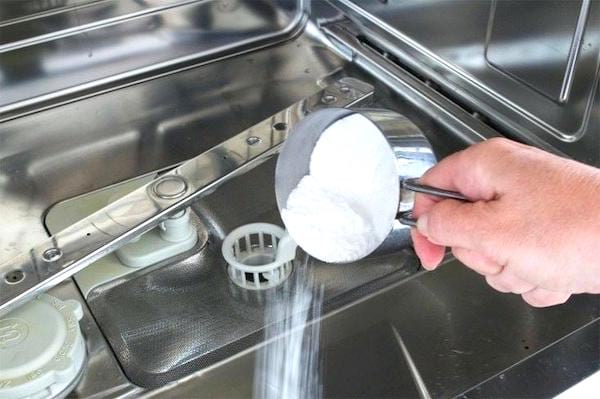Une main qui verse du bicarbonate de soude dans un lave-vaisselle à vide.