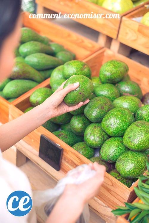 Des mains qui choisissent un avocat mûr dans un bac en bois rempli d'avocats.
