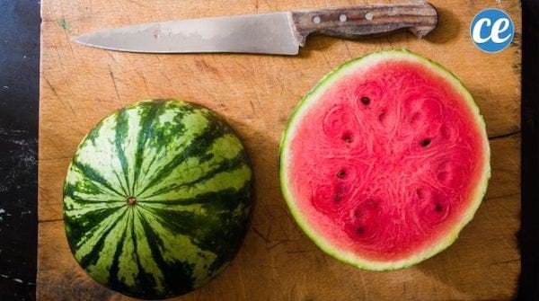 Un couteau long et une pastèque coupée en deux sur une planche en bois.