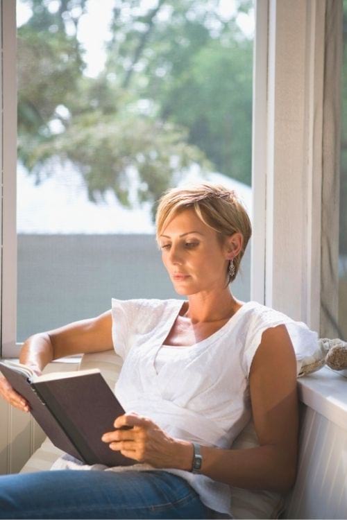 Une femme lit un livre à côté d'une fenêtre.