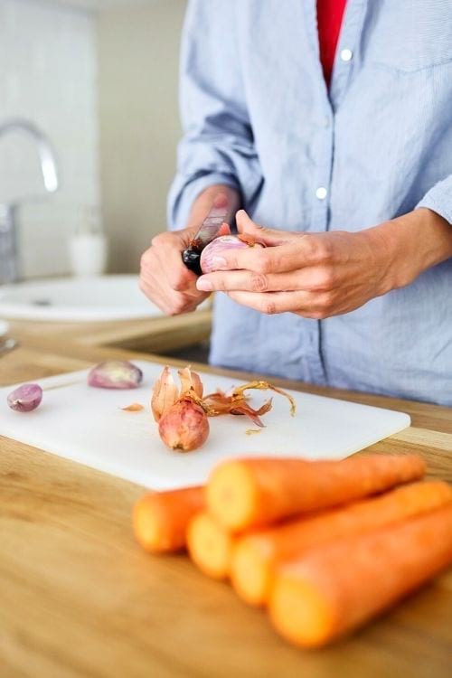 Des mains qui épluchent des carottes et des échalottes sur une planche à découper blanche.
