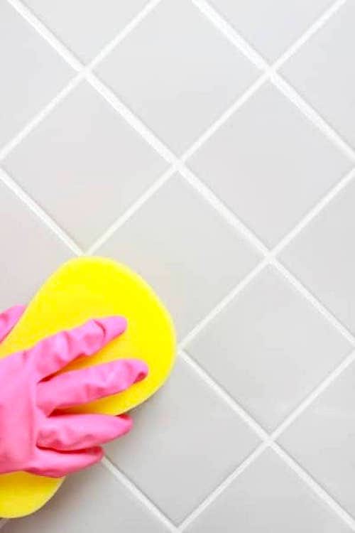 Une main dans un gant de nettoyage qui essuie des joints de carrelage avec une éponge.