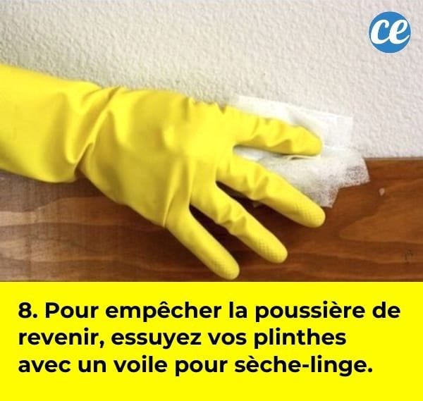 Une main dans un gant de ménage jaune qui nettoie une plinthe en bois avec un voile assouplissant pour sèche-linge.