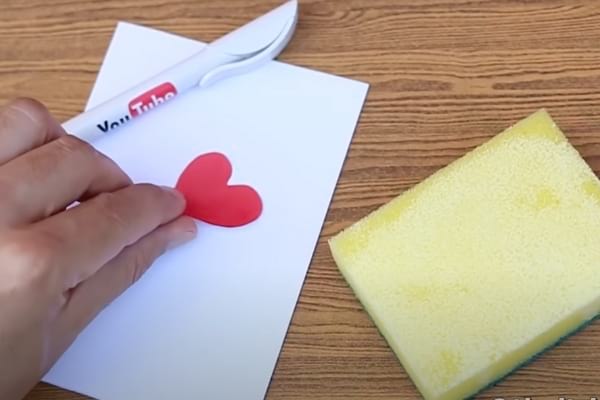 Petit coeur rouge collée sur une feuille blanche avec une éponge à proximité utilisée pour faciliter le collage