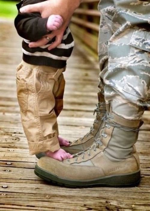 Un petit bébé qui est pieds nus sur les chaussures de son papa militaire