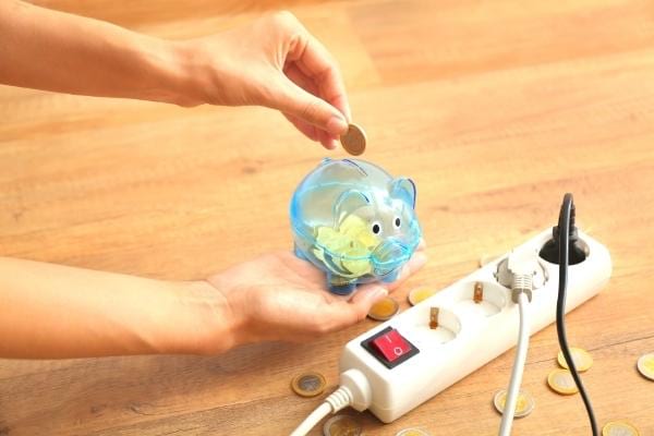 Eine Hand legt Pennys in ein Sparschwein neben einer Steckdosenleiste, um Strom zu sparen