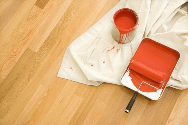 un pot de peinture rouge orangé avec un rouleau et une baille sur un drap blanc et parquet