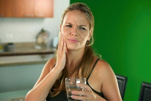 une femme avec un verre d'eau se tient la joue à cause d'un mal de dents