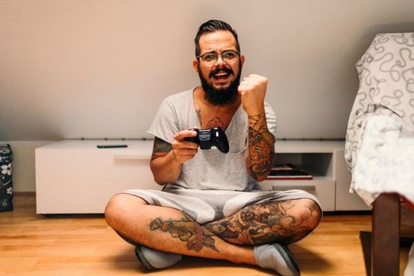 un homme barbus et tatoués assis sur le sol joue aux jeux vidéos
