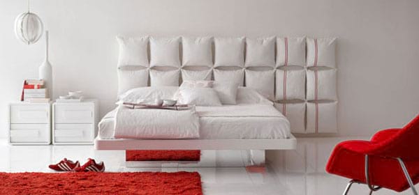 Un lit blanc avec plusieurs coussins derrière avec un siège et un tapis rouge devant le lit