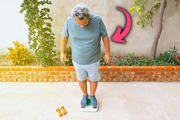 Un homme âgée habillé sur un pèse personne avec deux petits haltères jaune au sol à côté 