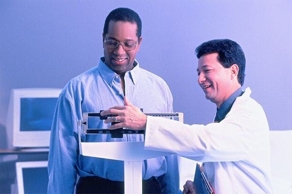 Un homme noir sur une balance chez un médecin qui se trouve juste à côté 