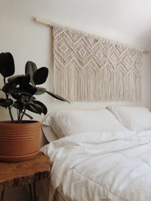 Un lit blanc avec une plante sur le côté avec une tête de lit avec du macramé suspendue