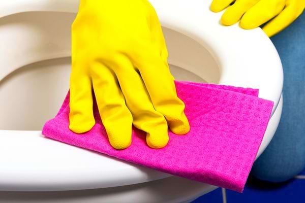 Eine Person, die mit gelben Handschuhen und einem violetten Mikrofasertuch ausgerüstet ist, reinigt eine Toilette