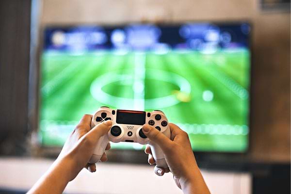 Une personne tenant une manette de Playstation 4 devant une télé avec un jeu de foot