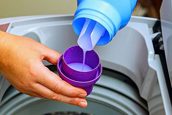 Une personne mettant de la lessive dans un bouchon violet au dessus d'une machine à laver 