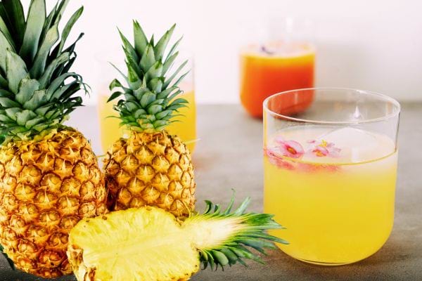 Trois ananas dont un qui est coupé en deux avec trois verres remplis d'une boisson de couleur jaune