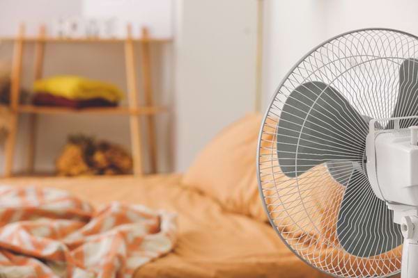 Une chambre avec un ventilateur devant un lit et une couette dérangée