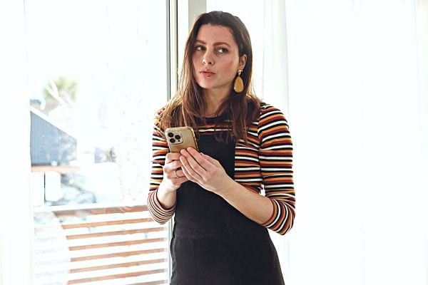 Une femme tenant son téléphone devant une fenêtre vêtu d'une salopette et d'un haut à barres