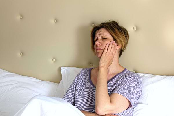 Une femme dans son lit se tenant la joue avec un air de souffrance 