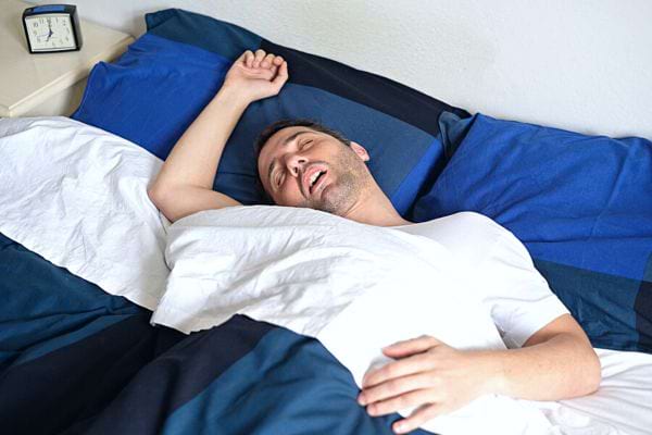 Un homme couché dans un lit avec de la literie bleue et la bouche ouverte et une petite horloge sur une table de chevet 