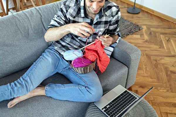Un homme pieds nus sur son canapé faisant du tricot devant un ordinateur 