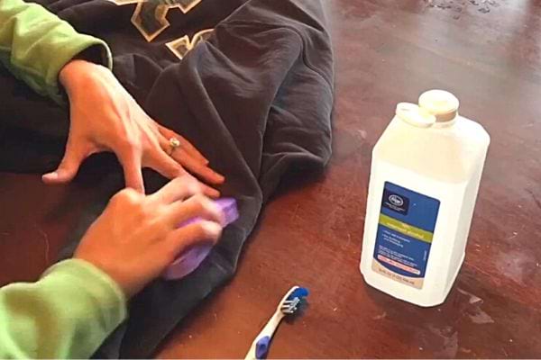 Eine Person reinigt eine schwarze Jacke mit einem violetten Tuch, einer Zahnbürste und einer weißen Flasche