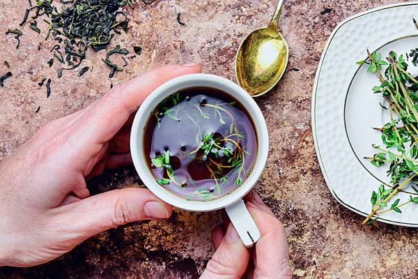 Tasse de tisane de thym, avec des feuilles de thym flottantes, tenue par deux mains sur un fond texturé.