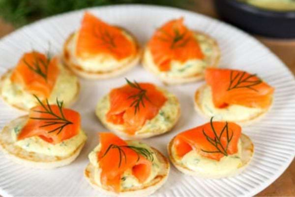 Petits blinis à l'aneth garnis de saumon fumé en forme de rose, disposés sur une assiette blanche élégante.