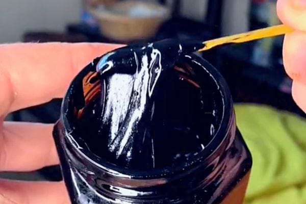 Résine de Shilajit noire étirée par un bâtonnet sur un pot tenu entre les doigts.