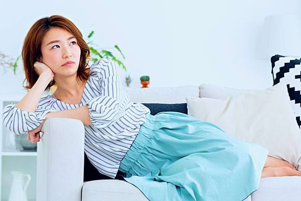 Une femme asiatique assise pensive sur un canapé, regardant au loin.
