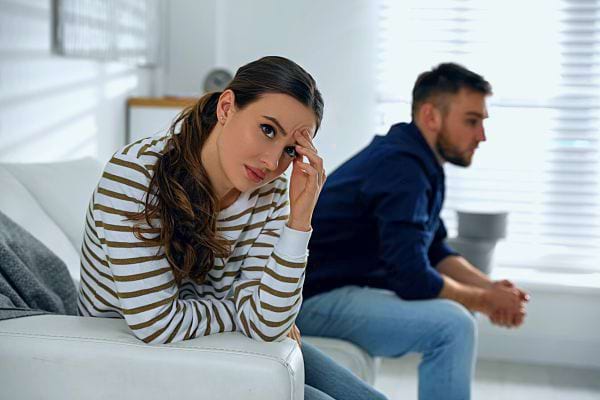 Une femme assise tient sa tête, l'air soucieuse, tandis qu'un homme est assis en arrière-plan, détourné.