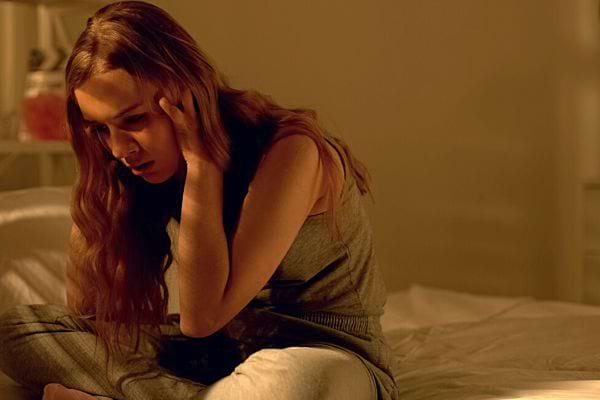 Une femme paraissant anxieuse, assise sur un lit, tête baissée, mains sur la tête.