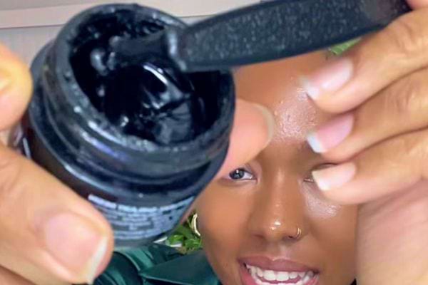 Pot de Shilajit ouvert, une spatule noire révélant la résine, devant un visage souriant.