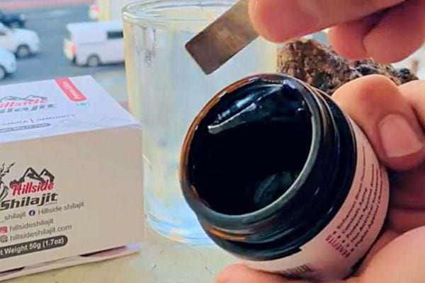 Main tenant un pot de "Natural Rems Himalayan Shilajit" ouvert sur fond extérieur.