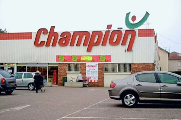Parking d'un supermarché Champion avec son enseigne rouge et une icône souriante verte au-dessus de l'entrée.