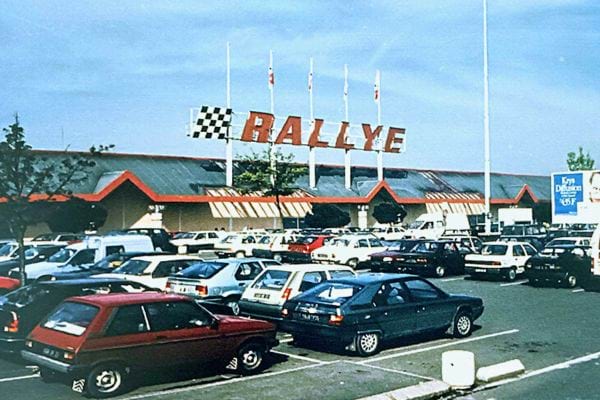 Parking du supermarché Rallye avec son enseigne à damier rouge et blanc et des drapeaux au-dessus.