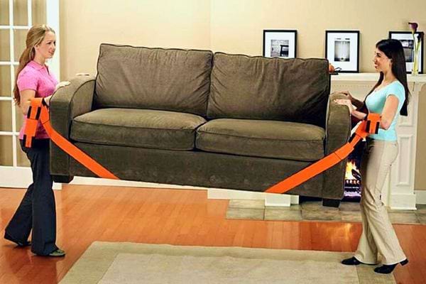 Deux femmes équipées de sangles de déménagement déplacent un grand canapé marron.
