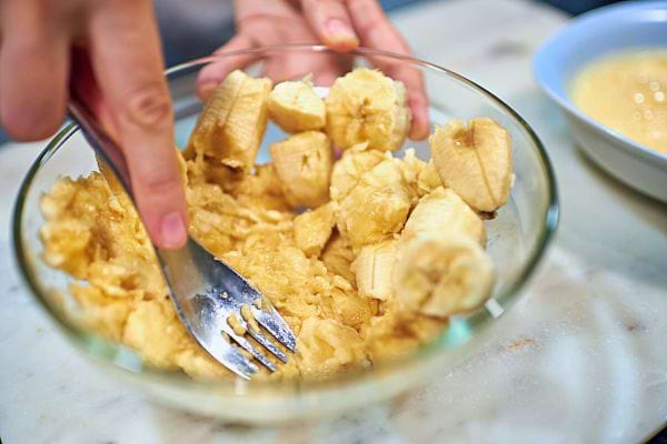 Mains écrasant des morceaux de banane dans un bol en verre avec une fourchette.