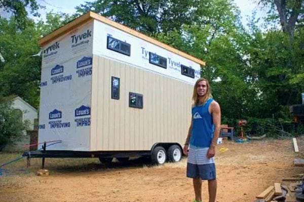 Étudiant devant sa maison mobile inachevée sur remorque.