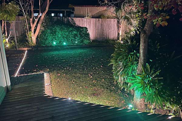 Jardin de nuit éclairé par des lumières automatiques, avec une pelouse et des arbustes visibles.