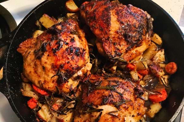 Deux cuisses de poulet assaisonnées dans une poêle en fonte avec des légumes rôtis.