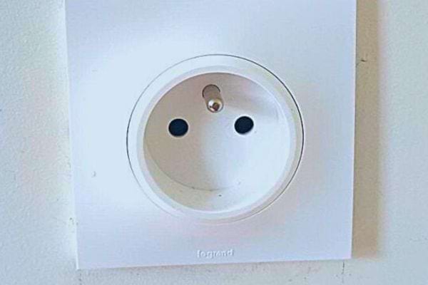 Prise électrique blanche simpliste avec deux trous et un trou de terre, évoquant un visage neutre.