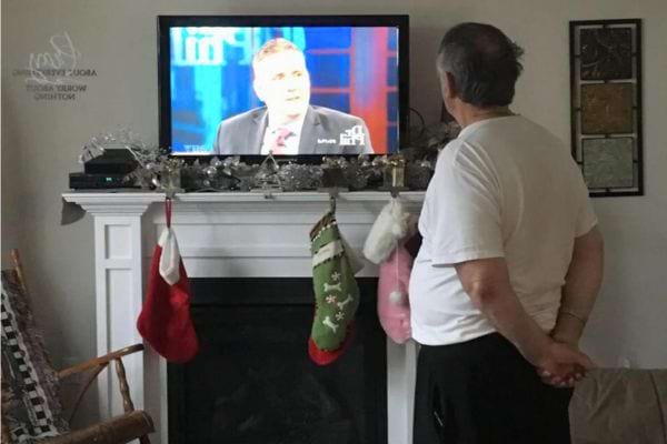 Un homme en t-shirt blanc regarde une télévision au-dessus d'une cheminée avec des chaussettes de Noël.