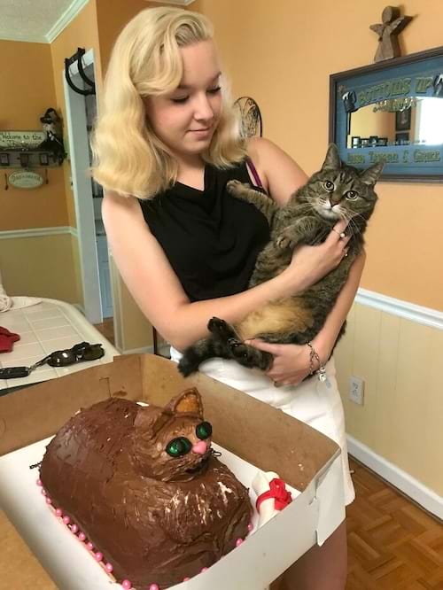 Femme tenant un chat à côté d'un gâteau qui lui ressemble, avec des yeux verts.