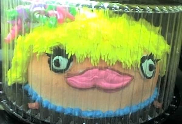 Gâteau rond avec visage aux cheveux fluo jaune, expression grotesque, très coloré.