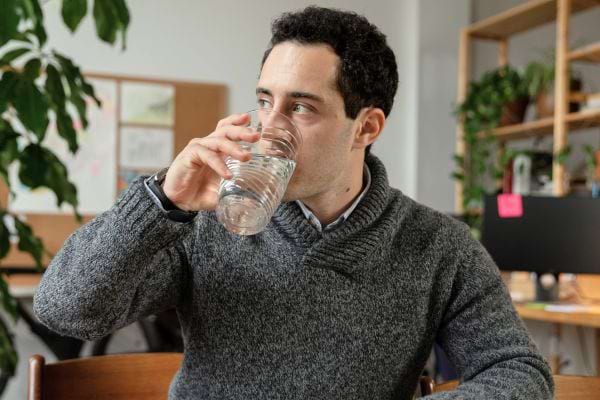 Homme buvant un verre d'eau dans un espace de bureau avec des plantes en arrière-plan.