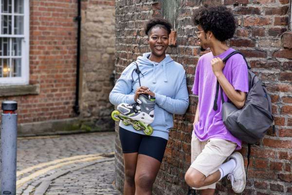 Deux amis, l'un avec des patins à roues alignées, partagent un moment de rire dans une rue ancienne.