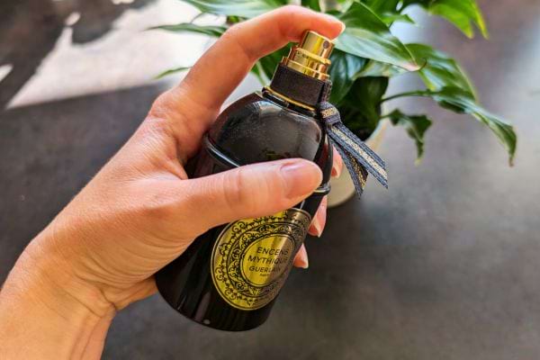 Main tenant un flacon de parfum noir devant des plantes.